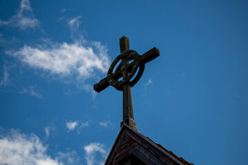 A weathered wooden Celtic cross stands atop a building against a bright blue sky with scattered...