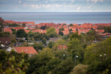 Obraz premium An elevated view captures a charming coastal town with red-roofed houses and lush green trees extending towards the calm blue ocean under a partly cloudy sky.
