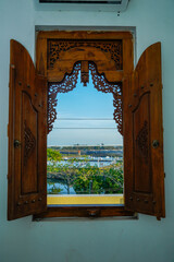 Wooden windows with carved motifs that open to reveal the outside view of water, trees and bright blue sky.