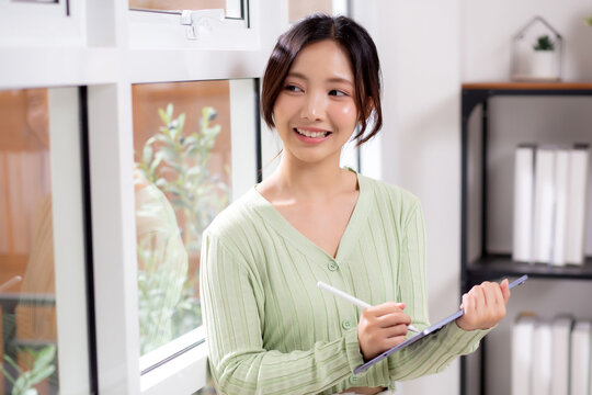 Young asian woman using tablet writing with inspiration and comfortable by window in living room at home office, cheerful woman standing working on digital tablet in workspace, business and lifestyle. - Powered by Adobe