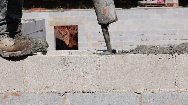 Construction worker stands atop cinder block wall, pouring freshly mixed concrete into gaps at building site.