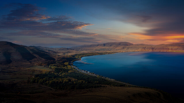 Panoramic view of Lake Sevan at sunset, with golden hills, calm blue water, and dramatic clouds under the colorful sky.