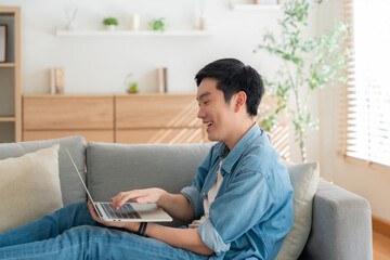 Fototapeta premium Young Asian man relaxing on comfortable couch, using laptop at home, happy boy chatting with friends in social network, spending weekend, watching movie, shopping online