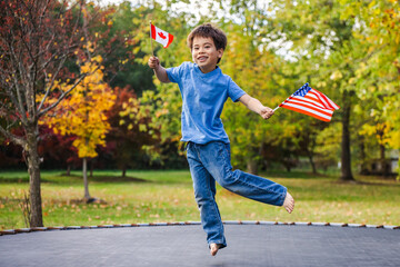 Happy boy jumping on trampoline holding American and Canadian flags in autumn park