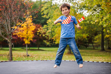 Happy boy jumping barefoot on trampoline holding American and Canadian flags during autumn day in a colorful park