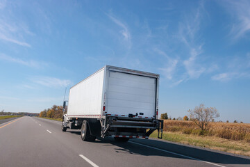 Angled view of the back of a white truck on a two-lane highway, driving on a clear sunny day with a blue sky and yellow fields in the background.