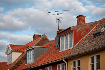 Bornholm - A row of colorful houses with red-tiled roofs, various dormer windows, chimneys, and an antenna stand against a cloudy blue sky.