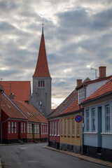 Obraz premium A narrow street winds through an old town lined with colorful houses, leading toward a historic church under a cloudy sky.