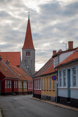 Obraz premium A picturesque street lined with vibrant houses leads to a striking church tower beneath a cloudy sky.