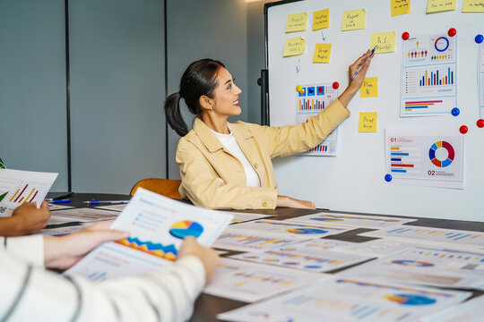 In a vibrant corporate setting, a dynamic young asian businesswoman presents important statistical insights to her engaged colleagues, analyzing data presented on a whiteboard in the office