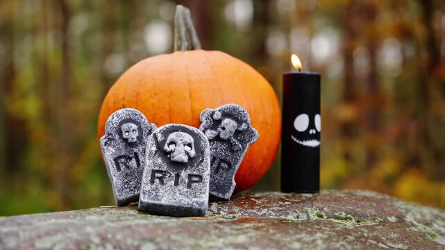 Skull gravestone decoration beside pumpkin on mossy forest floor, creepy halloween setup and flickering candle