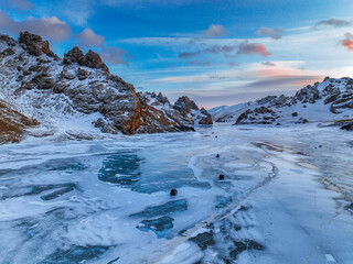 Wide view of the frozen Kel Suu Lake showing cracked blue ice and small dark rocks, set against snowy, rugged mountains at dusk