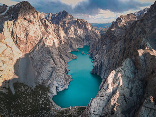 High-angle aerial view of the vibrant turquoise Kol Suu Lake nestled deep in a massive, rocky canyon under a dramatic, colorful sky