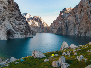 Ground-level view of the calm turquoise water of Kol Suu Lake, with small tents pitched among rocks and massive canyon walls