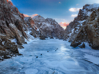 Stunning view of the frozen Kel Suu Lake and the surrounding steep, snow-dusted canyon walls under a colorful sunset or sunrise sky