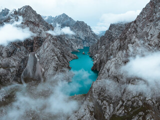 Dramatic aerial view of the turquoise Kol Suu Lake nestled deep within rugged canyon walls, partially obscured by low-hanging clouds and mist