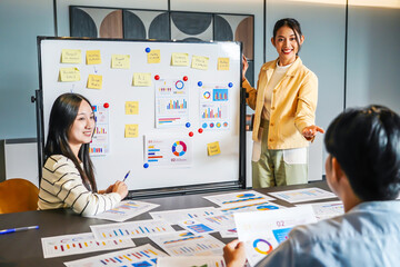 Young asian businesswoman conference speaker passionately explains business project strategy using a whiteboard, engaging a diverse team in collaborative brainstorming within a contemporary office
