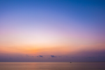 Background seascape dusk reveals a dramatic sky graduating from deep blue to pale orange and pink near the horizon. The smooth ocean reflects the colors, accompanied by the silhouette of a small boat.