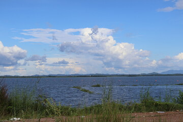 Fototapeta premium A serene lakeside view under a partly cloudy sky. In the foreground, there's an expanse of calm water reflecting the clouds above