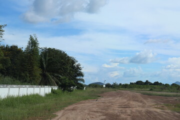 This is an image showing an open area with a paved road leading into it. The ground appears dry, and there are some structures or buildings on the far side of the field