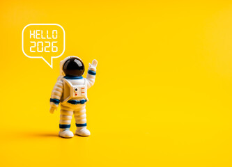 Cute astronaut figure waving with speech bubble “Hello 2026” on yellow background with copy space. Futuristic and minimal new year greeting concept for 2026. Creative space-themed celebration idea.