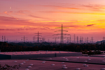 Ein spektakulärer Sonnenuntergang taucht eine Industrielandschaft in sattes, glühendes Orange und leuchtendes Magenta. Die Szene wird von einer Vielzahl von Hochspannungsmasten dominiert, deren Silhou