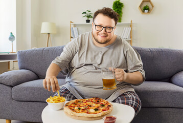 Overweight happy man eating fries and pizza, drinking beer at home. Funny hungry overweight guy in glasses sitting on sofa, holding mug and potato chips, unhealthy diet choice and obesity of person