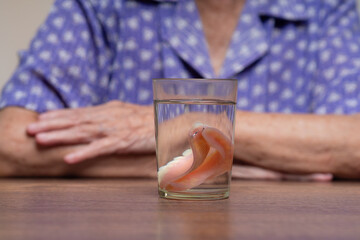 Dentures soaking in glass of water with elderly woman in background.