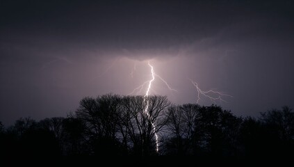 Striking lightning bolt illuminating rural night sky under dark clouds, with bare tree silhouettes