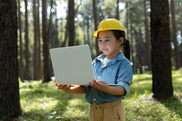 Child girl in a yellow safety helmet using a laptop outdoors.