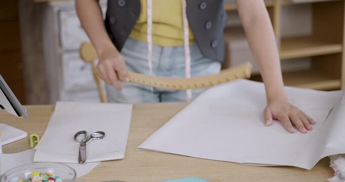 young asian female dressmaker measuring and marking pattern on fabric using pencil and ruler for creating handmade clothing inside fashion studio tailoring workspace with sewing tools on table