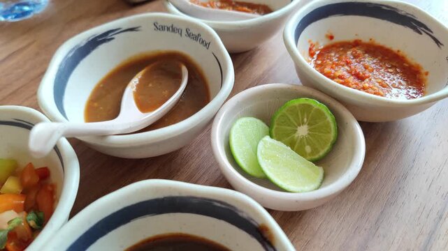 Close-up of traditional Indonesian seafood sauces and condiments served in ceramic bowls on a wooden table. The photo shows a variety of dipping sauces including sambal, soy sauce mix, and sliced lime
