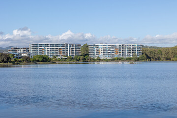 Several modern apartment buildings, two of equal height, line the shore of a lake and come with...