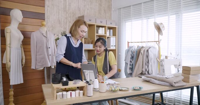 Asian fashion designers reviewing digital sketch and design ideas on tablet together in tailoring studio surrounded by sewing tools, handmade garments and fashion materials