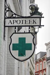 Fototapete Rund Apotheke An old pharmacy sign hangs on a white brick building. The sign says 'APOTEEK' and has a green cross, in a European city setting.  © Henk