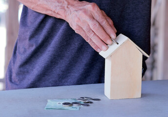 A man's hand is dropping coins into a wooden house-shaped piggy bank. On the table are coins and banknotes, representing the concept of saving money, financial planning, and investing for housing."