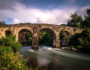 Fototapeta premium An old stone bridge with multiple arches gracefully spans a river. Long exposure creates blurred water motion, surrounded by greenery and a cloudy sky