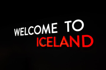 A bold neon sign reads WELCOME TO ICELAND on a deep black field, with strong even lighting and...