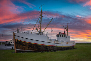 A weathered white trawler rests on a grassy embankment in Iceland at sunset. Rigging and wheelhouse show a historic workboat, wide angle view with golden hour light. © Aerial Film Studio