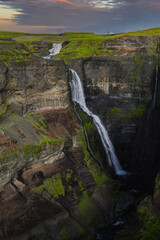 Multi tiered Glymur Waterfall plunges into a volcanic gorge in Hvalfjordur, Iceland at sunset. Warm pastel light contrasts dark cliffs and vivid green turf above.