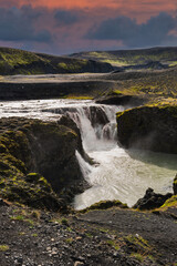 Compact multi tiered waterfall plunges through mossy lava canyon into milky glacial river in Iceland, with dark basalt, low clouds, and layered ridgelines at dusk.