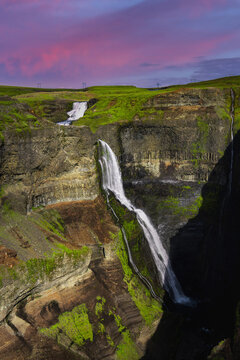 Haifoss and Granni waterfall pour over stratified basalt into a deep gorge in Thjorsardalur valley, Iceland. Magenta twilight light softens the dark ravine and green plateau.