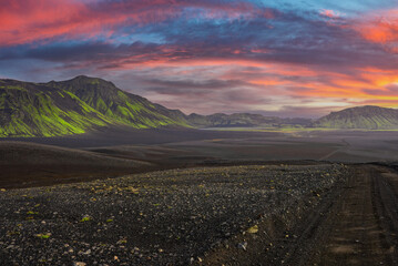 Moss lined volcanic mountains glow green over dark lava sands in Iceland. A gravel track leads into...