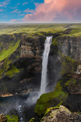 Haifoss plunges into a dark basalt canyon in Thjorsardalur, Iceland at golden hour, soft pink clouds and vivid green tundra framing mist rising from the pool below.