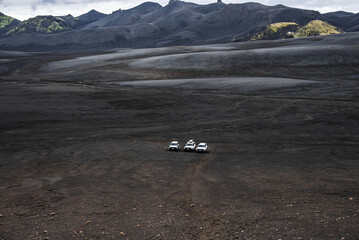 Three white 4x4 vehicles drive across black sand and ash in Iceland, wide and low view, soft...