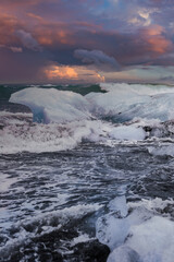 Jagged glacier ice rests on black sand as waves surge at Diamond Beach, Jokulsarlon, Iceland. Low golden light catches surf and ice under heavy evening clouds.