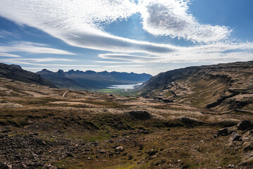 A wide valley leads to a distant fjord in eastern Iceland, with serrated ridges, mossy terrain, and a gravel road. Midday light defines rock textures and shimmering water.