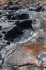 Close view of a geothermal mud field in Iceland, with cracked clay, dark viscous pools, rust hued deposits, and sulfur stains under diffused daylight, emphasizing volcanic activity.