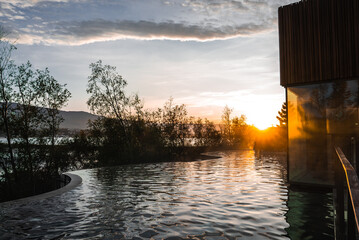 Steam rises from a curved geothermal pool beside a timber spa at dusk in Iceland. Birch trees, a calm lake, and low mountains reflect warm light under layered clouds.