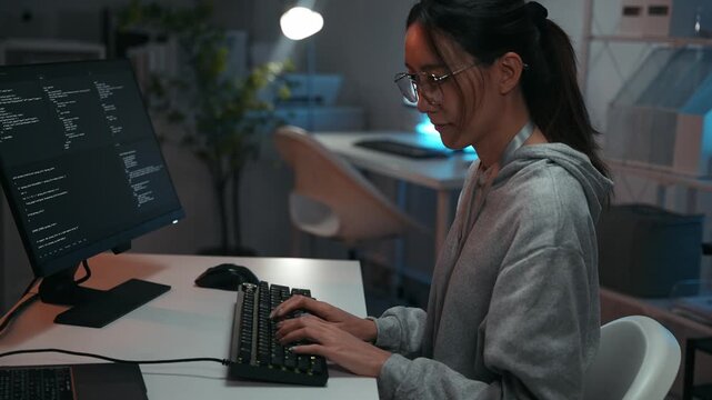 Focused Asian woman programmer working late at desk, typing on a keyboard with code on the computer screen. Developing complex software for data and cyber security in a dark office